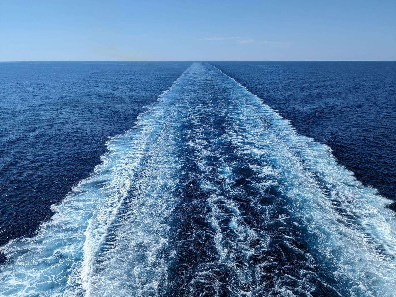 A ship's wake trailing across open blue ocean toward the horizon under a clear sky.