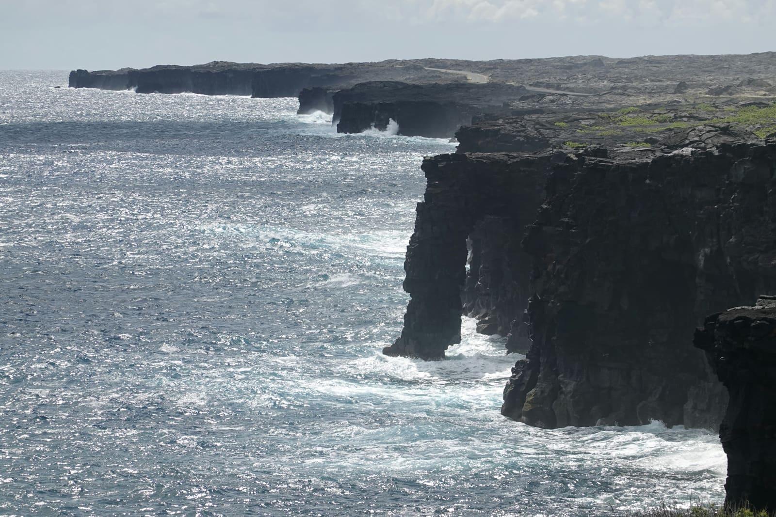 Black volcanic sea cliffs and rock arches along a Hawaii Island coastline, with waves breaking below and a paved road visible along the clifftop.