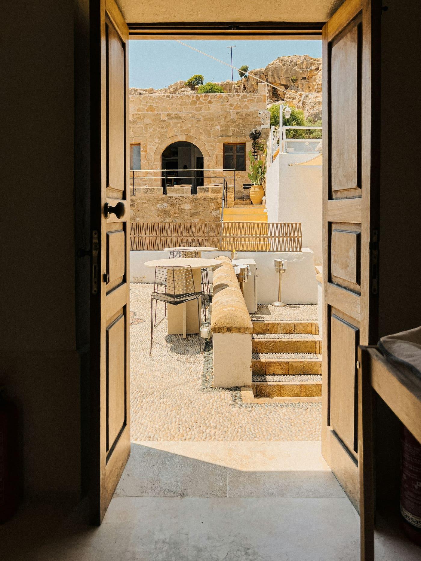 An open wooden double door leading to a sunlit stone courtyard with outdoor seating, a stepped walkway, and a rocky hillside beyond.